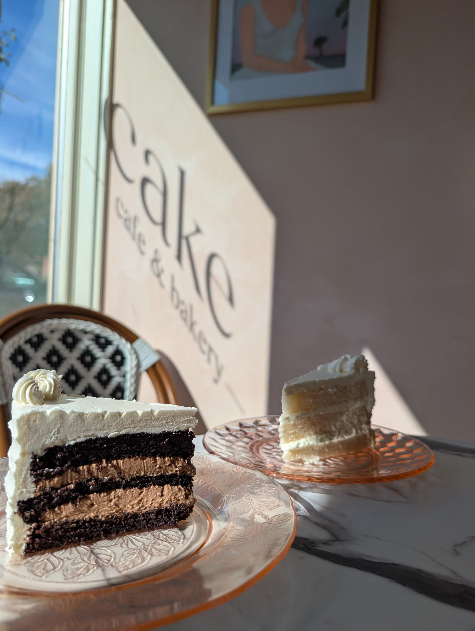 A chocolate layer slice and a vanilla slice on pink glass plates under the cake wall sign